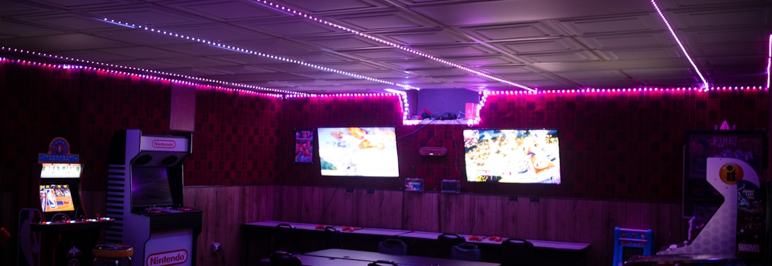 A dimly lit room with vibrant purple LED strip lights on the ceiling, featuring multiple gaming stations. There are two large screens showing video games, along with various arcade machines, including a Nintendo arcade cabinet. An air hockey table is positioned in the foreground.